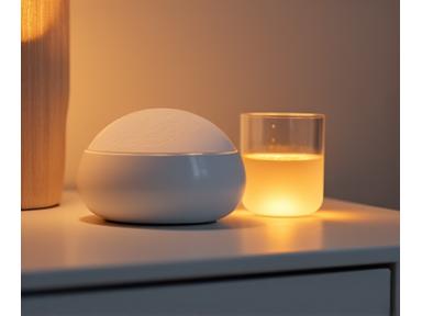 A small, elegant white noise machine emitting a soft glow on a nightstand, next to a glass of water, promoting a calm atmosphere.
